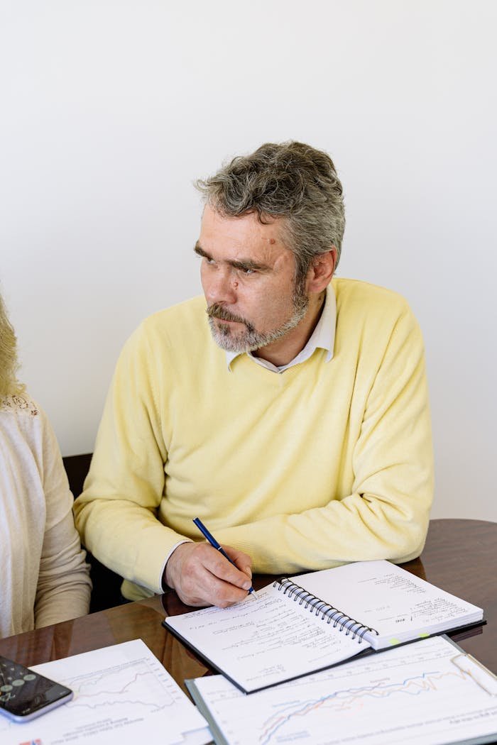 Home Elderly man in a yellow sweater writing with a pen in a notebook, seated at a desk.